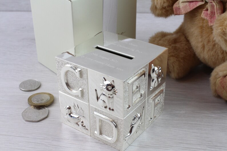 May include: Silver-plated baby block money box with a slot on the top. The box is decorated with letters, animals, and a design of a duck. The box is sitting on a wooden surface.