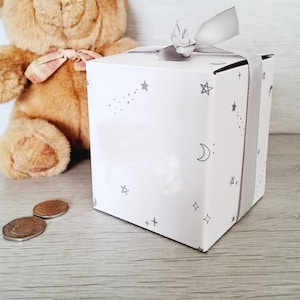 May include: A white gift box with a gray ribbon and a star and moon pattern. The box is sitting on a white surface next to a brown teddy bear.