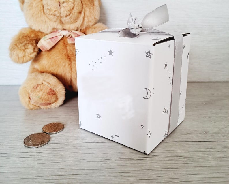 May include: A white gift box with a gray ribbon and a star and moon pattern. The box is sitting on a white surface next to a brown teddy bear.