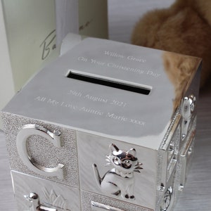 May include: Silver-colored baby block money box with a slot on the top. The box is engraved with the text "Willow Grace On Your Christening Day 29th August 2021 All My Love Auntie Marie xxxx".