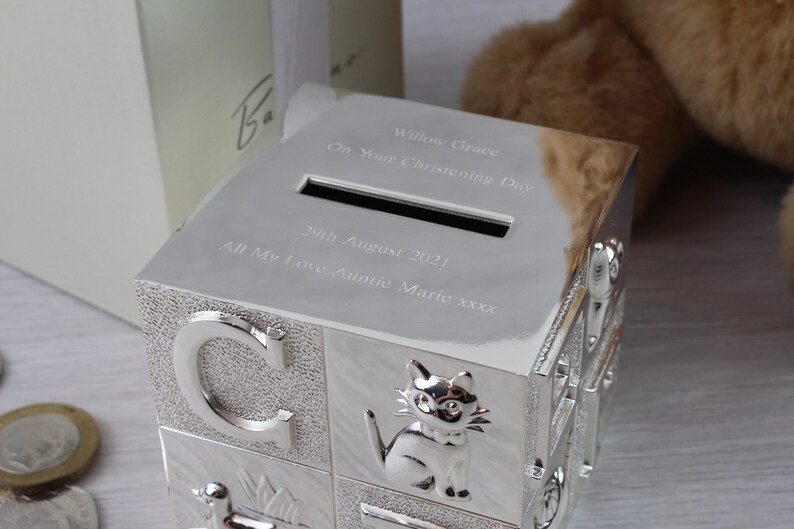 May include: Silver-colored baby block money box with a slot on the top. The box is engraved with the text "Willow Grace On Your Christening Day 29th August 2021 All My Love Auntie Marie xxxx".