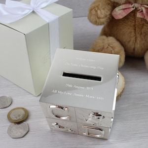 May include: Silver baby block money box with a slot for coins. The box is engraved with the text "Welcome Baby! It's Your Christening Day 9th August 2014 All My Love Auntie Mary xxx".