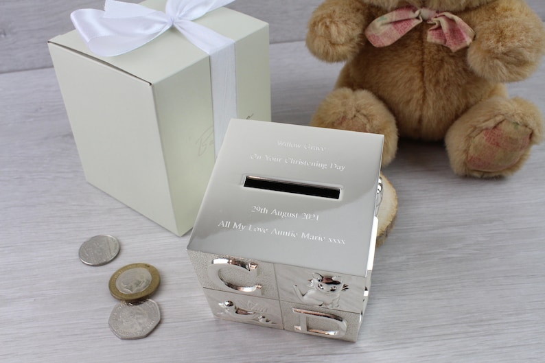 May include: Silver baby block money box with a slot for coins. The box is engraved with the text "Welcome Baby! It's Your Christening Day 9th August 2014 All My Love Auntie Mary xxx".