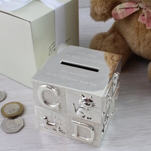 May include: Silver-plated alphabet block money box with a slot on the top. The box has a personalized inscription that reads "William George (The New & Little King) Born 29th August 2021 All My Firsts Auntie Marie xxx".