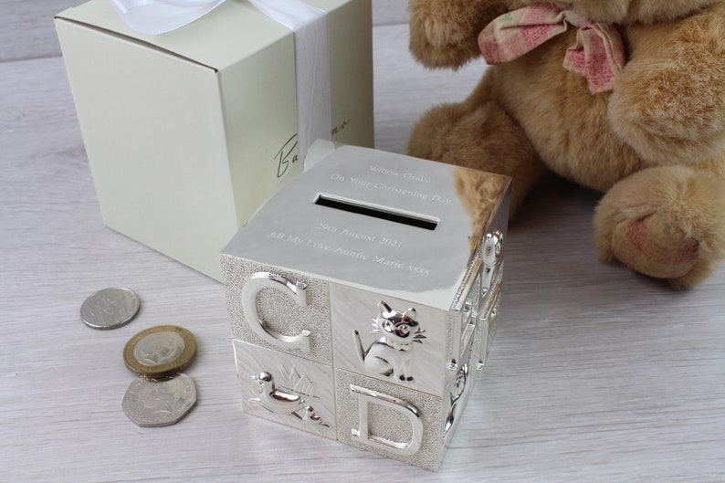 May include: Silver-plated alphabet block money box with a slot on the top. The box has a personalized inscription that reads "William George (The New & Little King) Born 29th August 2021 All My Firsts Auntie Marie xxx".