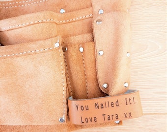 May include: A tan suede leather tool belt featuring multiple pockets, silver-coloured metal rivets, and a black adjustable strap. A leather loop attached to the side is embossed with the text "You Nailed It! Love Tara xx" in black lettering.