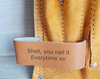 May include: A yellow suede leather tool belt featuring multiple pockets, white stitching, and silver metal rivets. A smooth tan leather loop is attached to the front, engraved with the black text: Shell, you nail it Everytime xx.