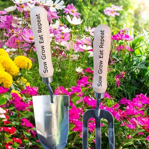 May include: Two stainless steel garden tools with wooden handles, a trowel and a three-tine cultivator, are displayed against a backdrop of colorful flowers. The trowel has a measurement scale in cm. The handles read "Sow Grow Eat Repeat".