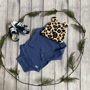 May include: A blue long-sleeved onesie, a pair of navy blue baby shoes, and a leopard print baby hat with ears on a wooden background.