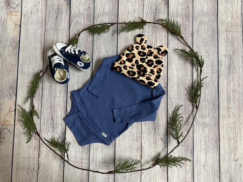May include: A blue long-sleeved onesie, a pair of navy blue baby shoes, and a leopard print baby hat with ears on a wooden background.