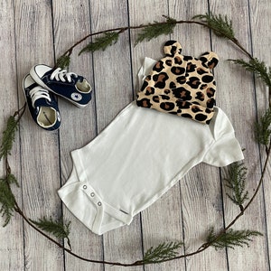 May include: A white baby onesie with a leopard print hat. The onesie has short sleeves and poppers at the crotch. The hat has a small bow on top.