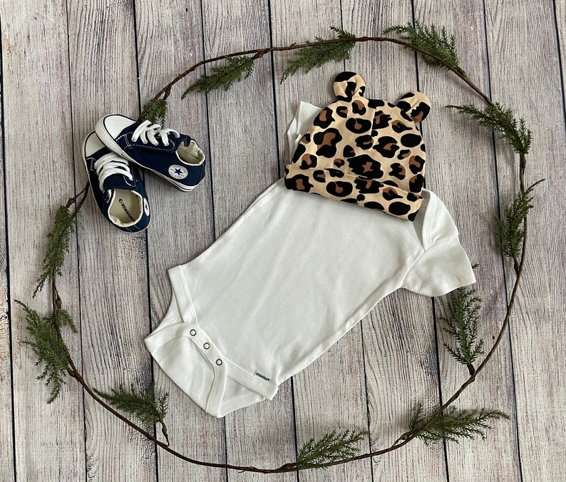 May include: A white baby onesie with a leopard print hat. The onesie has short sleeves and poppers at the crotch. The hat has a small bow on top.