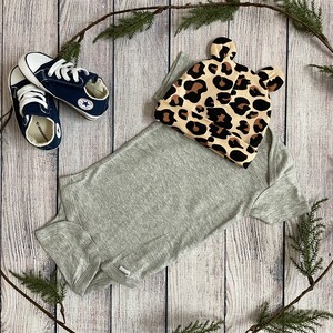 May include: A grey bodysuit with a leopard print hat featuring ears. The hat has brown and tan spots on a beige background. Two blue and white trainers are also in the image.