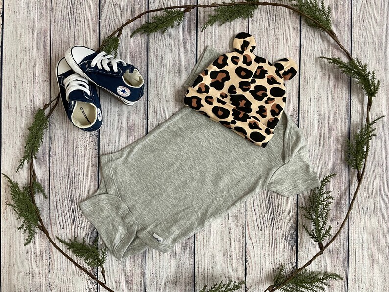 May include: A grey bodysuit with a leopard print hat featuring ears. The hat has brown and tan spots on a beige background. Two blue and white trainers are also in the image.