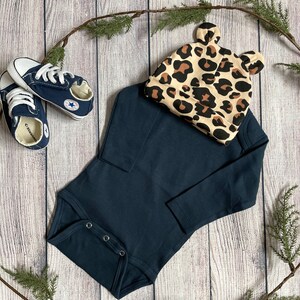 May include: A navy blue long-sleeved bodysuit with snap closures, a pair of blue canvas sneakers, and a leopard print hat with bear ears.