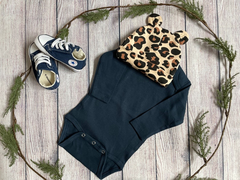 May include: A navy blue long-sleeved bodysuit with snap closures, a pair of blue canvas sneakers, and a leopard print hat with bear ears.