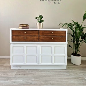 May include: A white and brown mid-century modern chest of drawers. It features four dark wood drawers with gold knobs above two white cabinet doors. The cabinet doors have a square design. The chest is from Blanche Furniture by MJ.