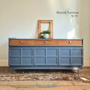 May include: A vintage-style sideboard with three drawers and three doors. The sideboard is painted a dark blue colour and has a wood top. The sideboard is sitting on hairpin legs. The text "Blanche Furniture by MJ" is visible on the front of the sideboard.