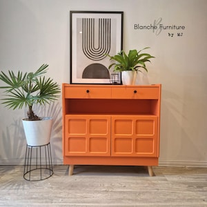 May include: A vibrant orange mid-century cabinet with two drawers and two doors, featuring a geometric pattern. The cabinet rests on wooden legs. A framed black and white artwork and two potted plants are displayed on top.
