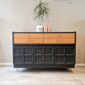 May include: A black mid-century modern sideboard with two drawers and three cabinet doors. The sideboard has copper-toned hardware and sits on hairpin legs. The sideboard is styled with a potted plant and two copper-toned bottles.