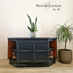 May include: A vintage black painted cabinet with two doors and two shelves. The cabinet has a hexagonal shape and is made of wood. The cabinet is sitting on a wooden floor. The text "Blanche Furniture by MJ" is visible on the wall behind the cabinet.