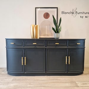 May include: A dark grey sideboard with gold handles and drawer pulls. The cabinet features a curved design and is topped with a gold vase, a potted plant, and a framed abstract art print. The text "Blanche Furniture by MJ" is visible.
