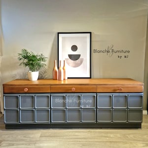 May include: A grey, mid-century modern sideboard with three drawers and a wood top. The sideboard has a geometric design with square compartments. The sideboard is decorated with copper drawer pulls. The sideboard is labelled "Blanche Furniture by MJ".