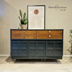 May include: A dark green mid-century modern sideboard with two drawers and three doors. The sideboard has gold-toned hardware and legs. The sideboard is sitting on a light wood floor. The text "Blanche Furniture by MJ" is visible on the front of the sideboard.