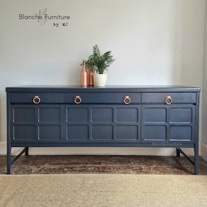 May include: A dark blue sideboard with copper hardware and a grid pattern on the doors. The sideboard has three drawers and three doors. It is sitting on a light brown rug in front of a white wall. The text "Blanche Furniture by MJ" is visible above the sideboard.