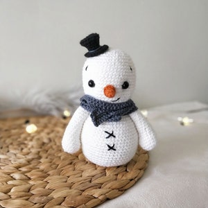 May include: A white crocheted snowman with a black top hat and a gray scarf. The snowman has black eyes, a small orange nose, and a stitched smile. The snowman is sitting on a woven straw mat.
