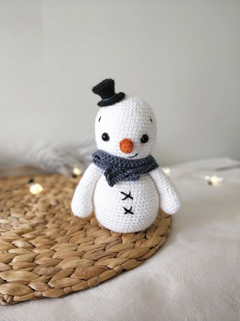 May include: A white crocheted snowman with a black top hat and a gray scarf. The snowman has black eyes, a small orange nose, and a stitched smile. The snowman is sitting on a woven straw mat.