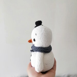 May include: A white crocheted snowman with a black top hat, orange nose, and gray scarf. The snowman is being held in a person's hand.