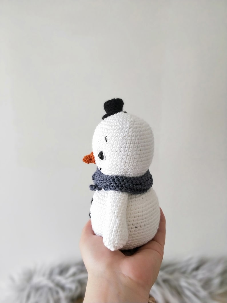 May include: A white crocheted snowman with a black top hat, orange nose, and gray scarf. The snowman is being held in a person's hand.