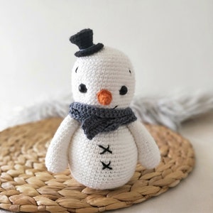 May include: A white crocheted snowman with a black top hat, gray scarf, and orange nose. The snowman is sitting on a woven straw mat.