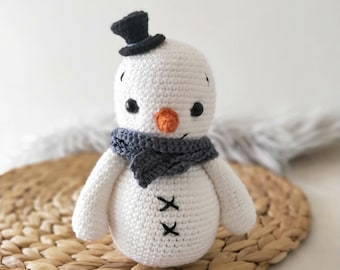 May include: A white crocheted snowman with a black top hat, gray scarf, and orange nose. The snowman is sitting on a woven straw mat.