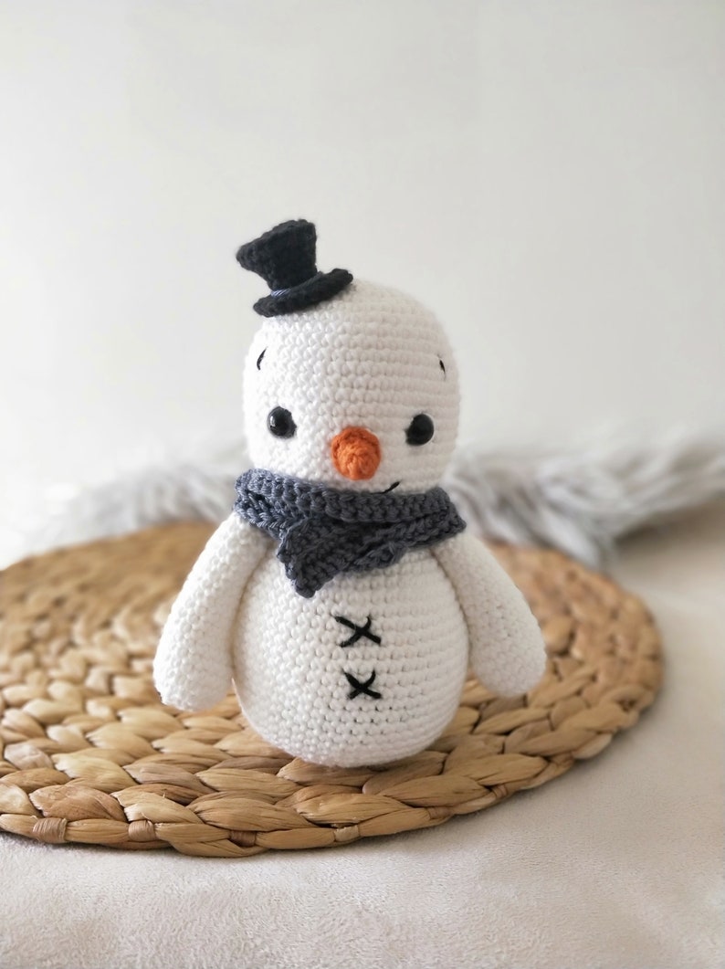 May include: A white crocheted snowman with a black top hat, gray scarf, and orange nose. The snowman is sitting on a woven straw mat.