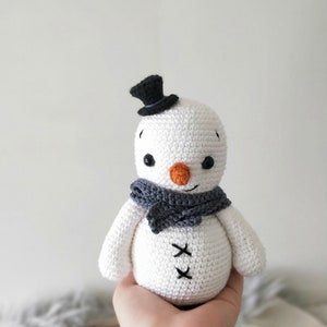 May include: A white crocheted snowman with a black top hat, gray scarf, and orange nose. The snowman has black eyes and a stitched smile.