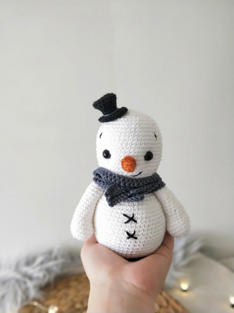 May include: A white crocheted snowman with a black top hat, gray scarf, and orange nose. The snowman has black eyes and a stitched smile.