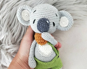 May include: A crocheted koala plush toy with gray fur, white ears, and a gray nose. The koala is wearing a green and brown outfit.