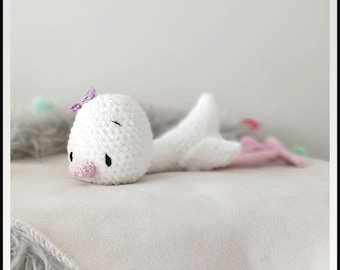 May include: A white crocheted stuffed animal shaped like a whale with a purple bow on its head. The whale is lying on a white fluffy surface.
