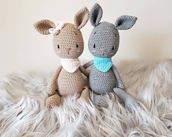 May include: Two crocheted stuffed animals, one brown and one gray, sitting on a fluffy white rug. Both animals have long ears and are wearing crocheted bibs.