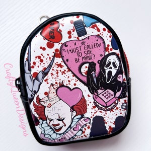 May include: A white and black mini backpack with a red and pink horror movie themed print. The print features characters from the movies "It", "Scream", and "Friday the 13th". The backpack has a zipper closure and a small handle on the top.