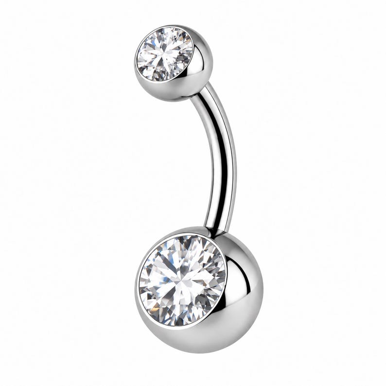 May include: A curved barbell belly button ring featuring a polished silver-tone metal finish. The jewelry includes a smaller top ball and a larger bottom ball, each set with a single clear, round-cut faceted gemstone that reflects light.