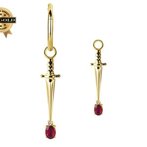 Op de afbeelding: Een paar goudkleurige oorbellen, elk met een dolk-hanger en een rode edelsteen. Eén oorbel is een creool, de andere is een enkele hanger. De afbeelding bevat een gouden zegel met de tekst "18K GOLD CERTIFIED".