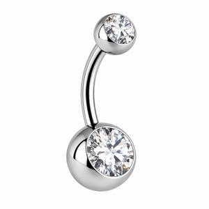 May include: A curved barbell navel ring made of polished silver-toned metal. The piece features two round clear faceted stones set into spherical ends, with a larger stone at the bottom and a smaller stone at the top of the curved bar.