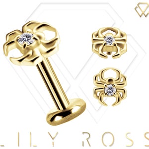 May include: Gold-toned spider body jewelry with a clear gemstone. The spider is shown in three different views: a close-up of the spider, a side view of the spider, and a view of the spider attached to a post.