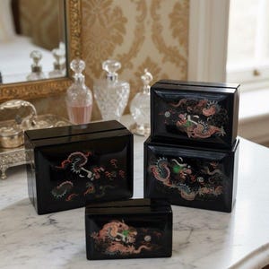 May include: Four black lacquered boxes with dragon designs in red, green, and gold. The boxes are rectangular with a glossy finish, arranged on a marble surface. The boxes vary in size, suggesting a set.
