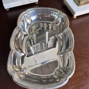 May include: A polished silver-tone metal tray with a scalloped, rounded rectangular shape. The tray features a smooth, reflective surface with curved, indented sides and a raised rim, creating a deep basin for holding small items or decorative objects.