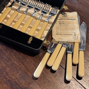 May include: A set of silver-toned forks and knives with cream-colored handles, presented in a black velvet-lined case. The set includes a paper certificate of guarantee with printed text, detailing the quality and craftsmanship of the cutlery.