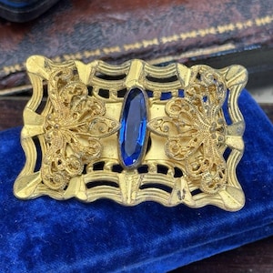 May include: A vintage gold-toned brooch with an oval, deep blue gemstone at its center. The rectangular brooch features ornate filigree and scrollwork designs. The brooch is displayed on a blue velvet surface.